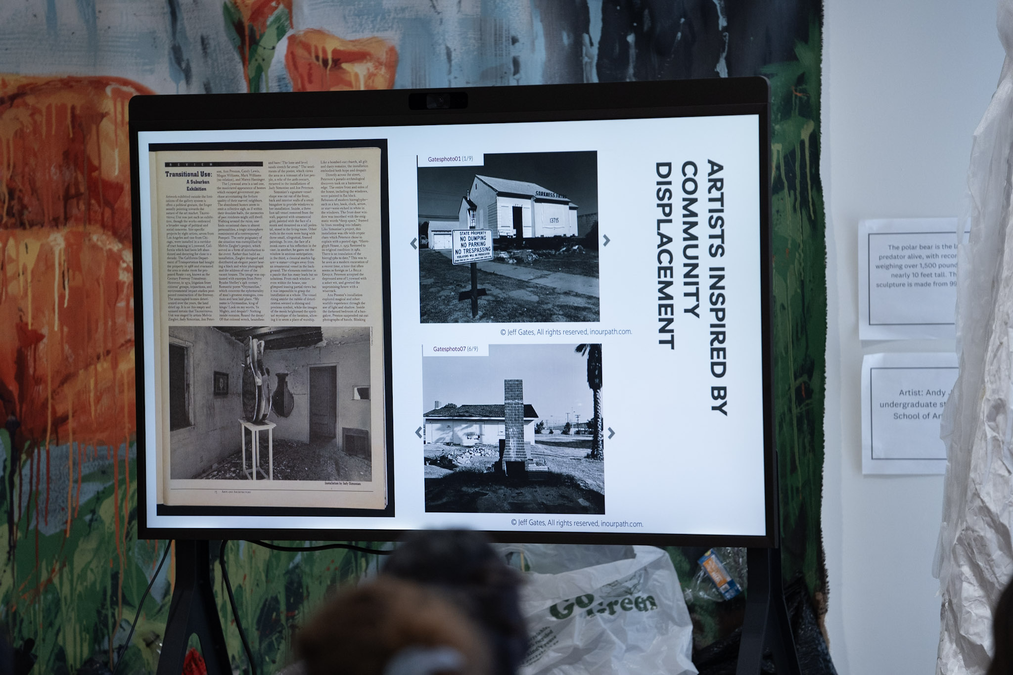 a monitor shows a slide titled "Artists Inspired by Community Displacement" displays newspaper clippings and photographs related to the construction of Los Angeles's Century Freeway