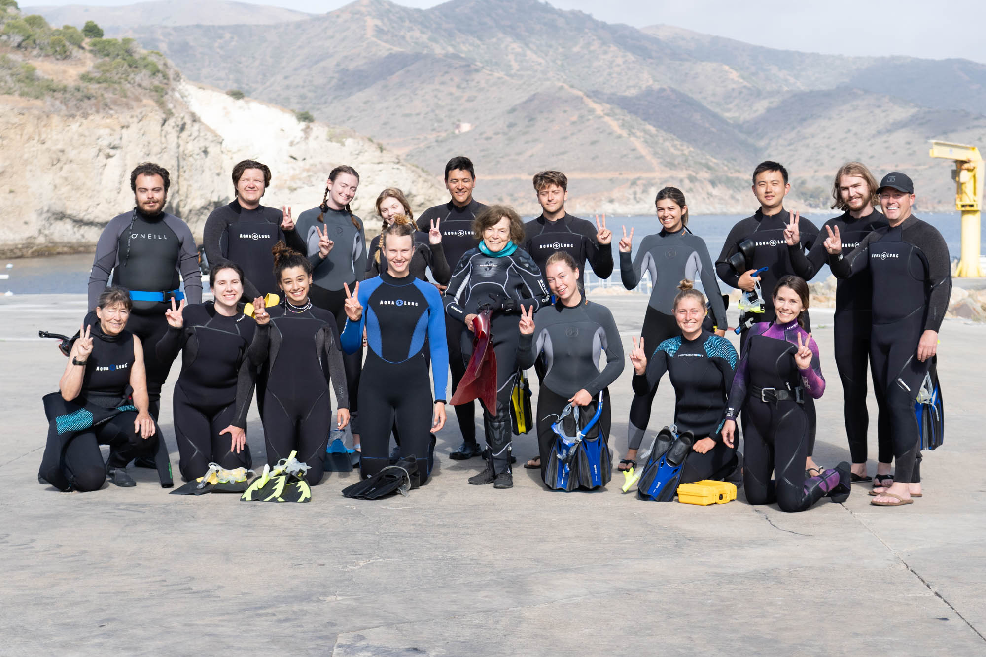 a group of people in wetsuits stands on a concrete dock with water and hills in the background