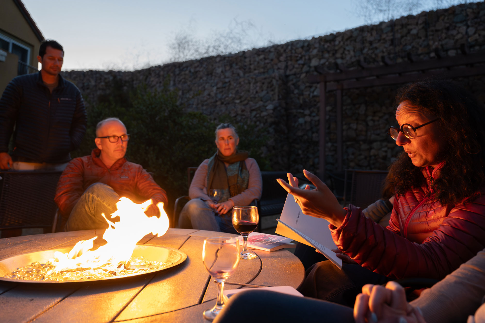 a woman wearing glasses gestures with her hand as she reads a book of poetry to colleagues around a campfire