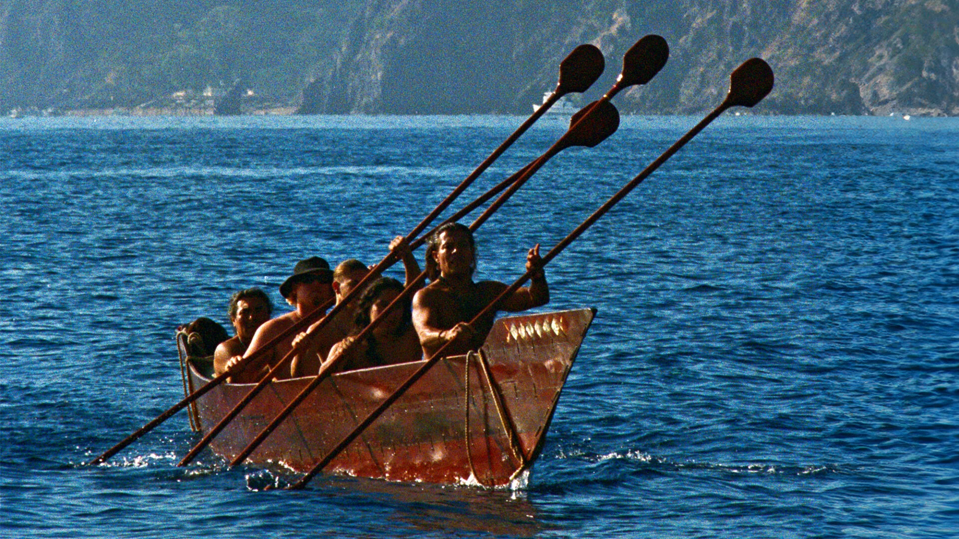 five people on a brown canoe in water hold paddles
