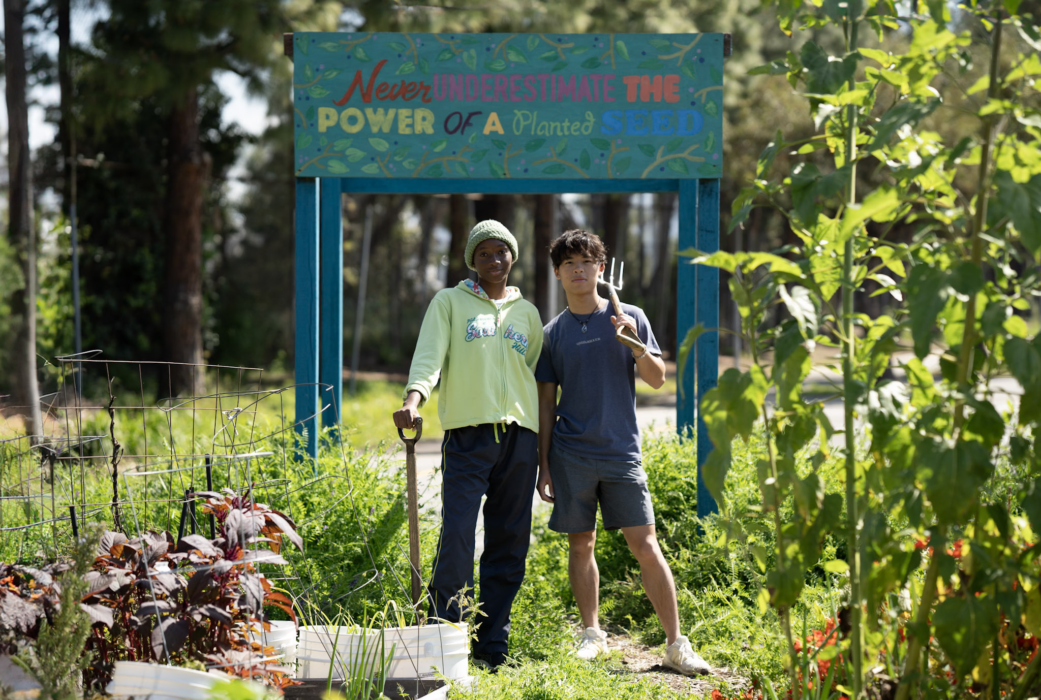 two student interns hold gardening equipment while posing in front of a sign that reads "never underestimate the power of a planet seed" in a vibrant garden